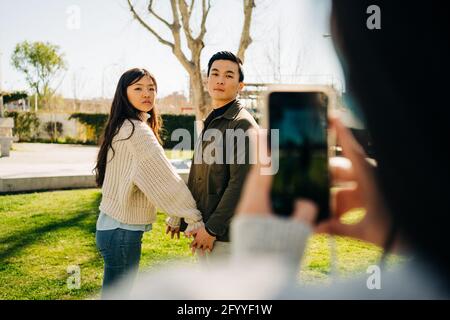 Crop unrecognizable Woman taking photo on smartphone of young Asian couple holding hands while standing on lawn in city park Stock Photo