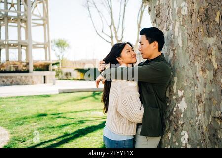 Happy young ethnic couple in casual clothes hugging and looking at each other standing near tree during romantic date in park Stock Photo