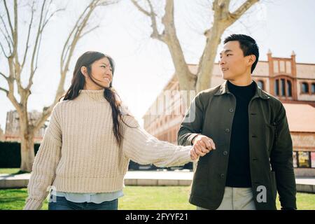 Happy young ethnic couple in stylish outfits holding hands while walking together in city park on sunny day Stock Photo
