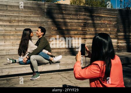 Side view of unrecognizable Woman using smartphone while taking photo of romantic young Asian couple hugging on wooden bench in city park Stock Photo