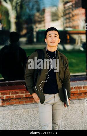 Positive young ethnic male millennial in stylish outfit smiling while enjoying favorite song in earphones walking on city street with laptop in hand Stock Photo