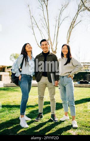 Full length of confident young Asian male and female students in trendy outfits smiling and looking at camera while standing on green lawn on sunny da Stock Photo