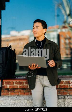 Thoughtful young Asian male freelancer in casual clothes looking away while working remotely on laptop standing near glass mirror building Stock Photo