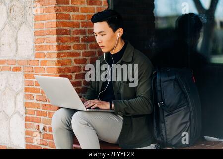 Thoughtful young Asian male freelancer in casual clothes looking at screen while working remotely on laptop sitting on bench near brick building with Stock Photo