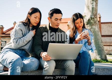 Young stylish male and female students with dark hair in casual clothes sitting on bench in campus and preparing for exams using laptop and smartphone Stock Photo