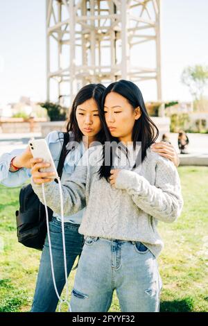 Serious young ethnic female friends cuddling while watching video on mobile phone standing on lawn in park near anonymous man Stock Photo