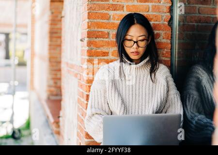 Stylish young Asian female remote worker in casual clothes and eyeglasses using laptop while sitting on bench near brick building