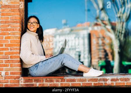 Stylish happy young Asian female remote worker in casual clothes and eyeglasses using laptop while sitting on bench near brick building