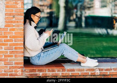 Side view of young Asian female freelancer in stylish sweater and eyeglasses smiling and browsing on message while working remotely on laptop on city Stock Photo