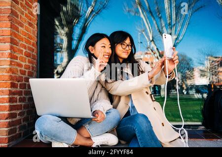 Cheerful young Asian female best friends with long dark hair in stylish outfits laughing while taking selfie on smartphone during work on laptop sitti Stock Photo