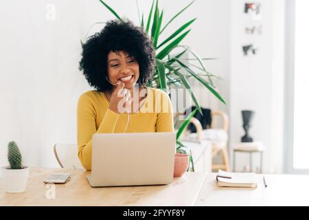Positive African American female smiling and working at table with netbook while using earphones having important phone call on smartphone Stock Photo