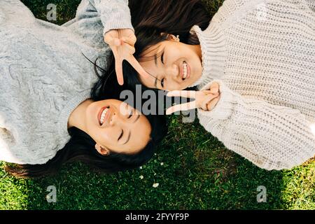 Top view of delighted young ethnic female best friends in stylish sweaters smiling and showing peace gesture while lying together on grassy lawn on su Stock Photo