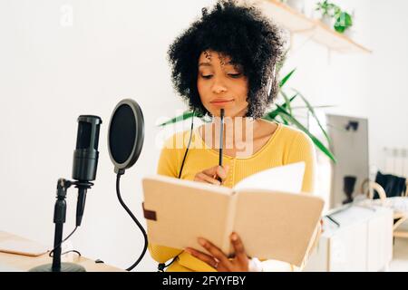 Thoughtful young ethnic female employee with dark Afro hair in casual clothes and headphones sitting at table with microphone and reading notes while Stock Photo