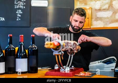 Bearded professional ethnic cutter preparing tasty jamon in restaurant with bottles of wine Stock Photo