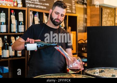Focused man sommelier with beard and moustache in black apron standing at bar counter and pouring bottle of red wine into glass decanter in restaurant Stock Photo