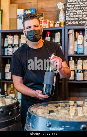 Serious bearded barman with dark hair in black apron standing at counter and demonstrating bottle of red wine while working in restaurant Stock Photo