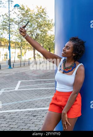 Full length positive African American female in summer wear taking selfie on mobile phone while standing on city street Stock Photo