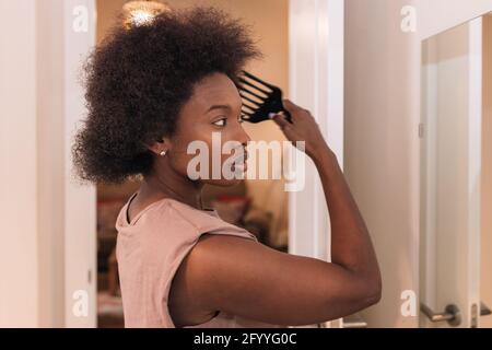 Side view of crop concentrated confident young African American female in casual clothes standing in front of mirror and brushing curly dark hair at h Stock Photo