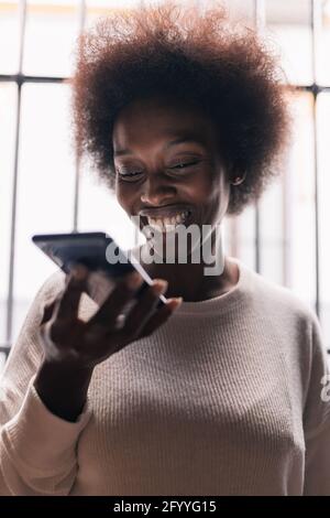 Joyful young African American female with curly dark hair smiling and recording voice message on mobile phone at home Stock Photo