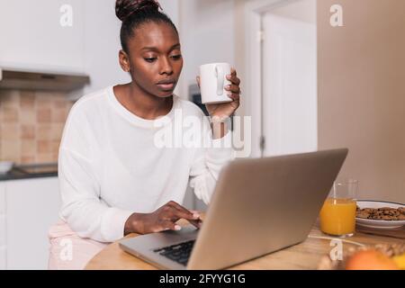 Side view of cheerful young black female freelancer in casual clothes smiling while typing on laptop during breakfast with coffee and fresh fruits at Stock Photo