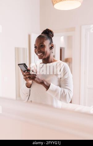 Happy young African American lady in casual clothes smiling while enjoying music in earphones with smartphone in hands Stock Photo