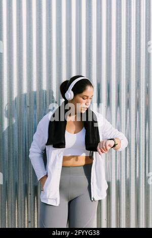 Active young ethnic female athlete in sportswear and headphones checking data on smartwatch while resting near metal wall during outdoor workout on su Stock Photo