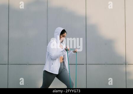 Side view of motivated young ethnic female athlete in sportswear doing exercise with resistance band during outdoor training on street Stock Photo
