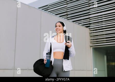 Positive young ethnic female athlete in sportswear smiling while messaging on mobile phone standing near metal wall on city street after workout Stock Photo