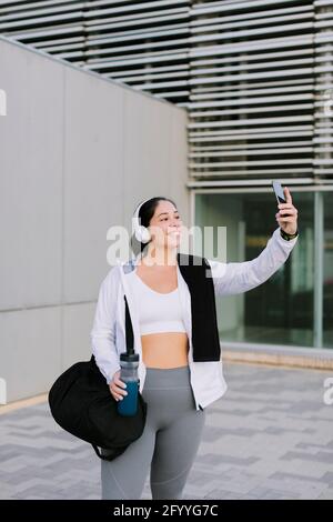 Cheerful young ethnic lady in sportswear and headphone smiling while taking selfie on smartphone after workout in city Stock Photo