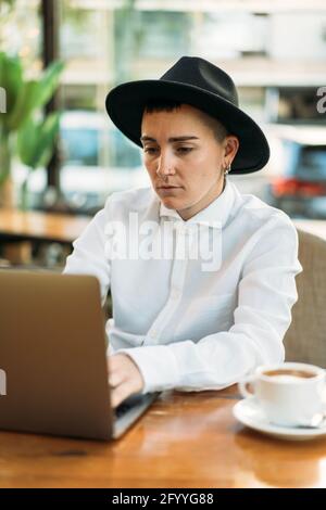 Concentrated masculine Woman wearing stylish hat and white shirt working on laptop while working remotely Stock Photo