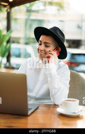 Stylish happy female freelancer having phone call while working in cafe with laptop with cup of coffee Stock Photo