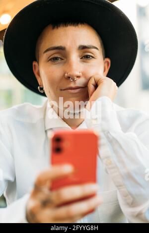 Serious masculine Woman wearing white shirt and hat with mobile phone sitting in cafe and thoughtfully looking away Stock Photo