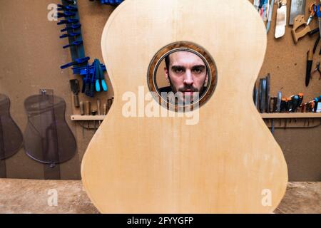 Crop serious male luthier looking at camera through sound hole of classical musical instrument in workshop Stock Photo