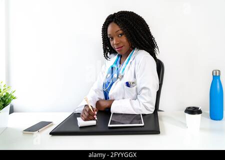 Positive black with Afro hairstyle wearing medical uniform sitting at table with tablet and writing prescription in notebook during work in modern cli Stock Photo
