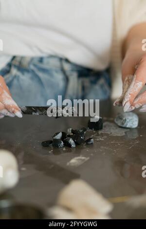 Crop anonymous female jewelry designer with dirty hands cutting clay while working in craftsmanship Stock Photo