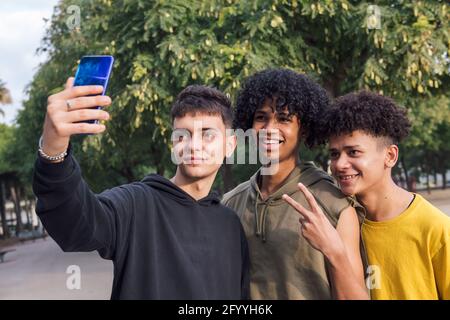 Delighted black male teens standing under tree and taking self portrait on mobile phone in park Stock Photo
