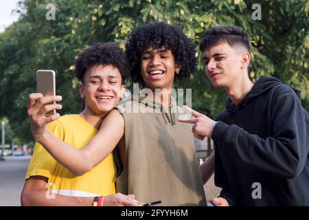Delighted black male teens standing under tree and taking self portrait on mobile phone in park Stock Photo