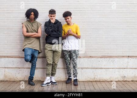 Full body of multiethnic black man with folded hands standing near friend touching chin close to ethnic teen against brick wall Stock Photo