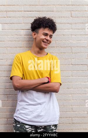 Cheerful African American teenage male in casual clothes smiling happily standing against brick wall and looking away Stock Photo