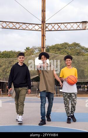 Confident multiracial male teens with basketball and skateboards standing together against sports net in park Stock Photo