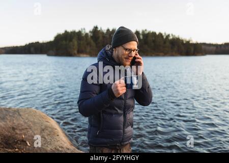 Young bearded male hiker in warm clothes and eyeglasses drinking hot beverage and talking on smartphone while standing near lake during trekking Stock Photo