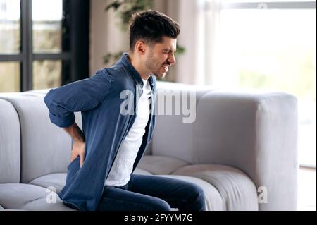 Sad upset young adult caucasian bearded man, in casual clothes, sitting on the sofa in the living room, massaging his back, suffering from back pain, suffering from arthritis, sciatica Stock Photo