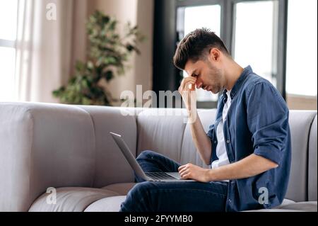 Upset tired young caucasian bearded man sitting on sofa in living room, in casual clothes, using laptop, student in stress, overworked with study and homework, rubs eyes, needs rest Stock Photo