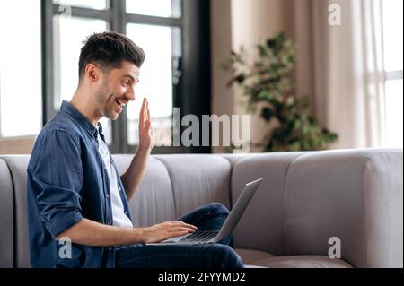 Happy friendly young caucasian bearded man sitting on a sofa, wearing stylish denim clothes, using a laptop, chatting with friends or family via video conference, waving his hand, smiling Stock Photo
