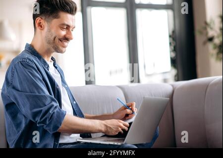 Smart confident young caucasian man sitting on the couch, in casual clothes, student doing online distance learning, freelancer works distantly, taking notes, looking at the screen, smiling Stock Photo