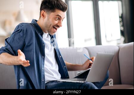 Sociable, friendly, emotional handsome caucasian guy sitting on the sofa in casual clothes, student talking during an online seminar, distance learning, group brainstorming, looking at screen, smiling Stock Photo