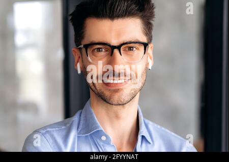 Close-up portrait of a handsome intelligent successful confident young adult man, ceo, wearing glasses and wireless headphones, wearing casual clothes, looking at camera, friendly smiling Stock Photo