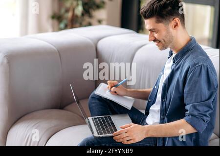 Smart confident young caucasian man sitting on the couch, in casual clothes, student doing online distance learning, freelancer works distantly, taking notes, looking at the screen, smiling Stock Photo