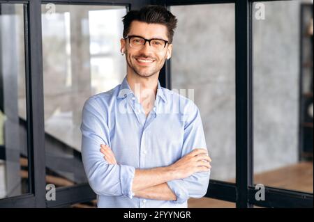 Portrait successful confident smart young adult business man or manager wearing glasses and headphones, wearing stylish clothes, standing in office, looking at camera, arms crossed, friendly smiling Stock Photo