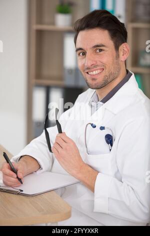 male doctor sat at desk writing on clipboard Stock Photo
